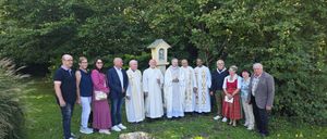 Eine Gruppe von Menschen, einschließlich Geistlicher und Laien, steht draußen in einem grünen Bereich, mit Bäumen und einer kleinen Kapelle im Hintergrund.