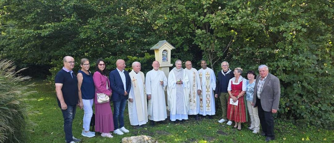 Eine Gruppe von Menschen, einschließlich Geistlicher und Laien, steht draußen in einem grünen Bereich, mit Bäumen und einer kleinen Kapelle im Hintergrund.