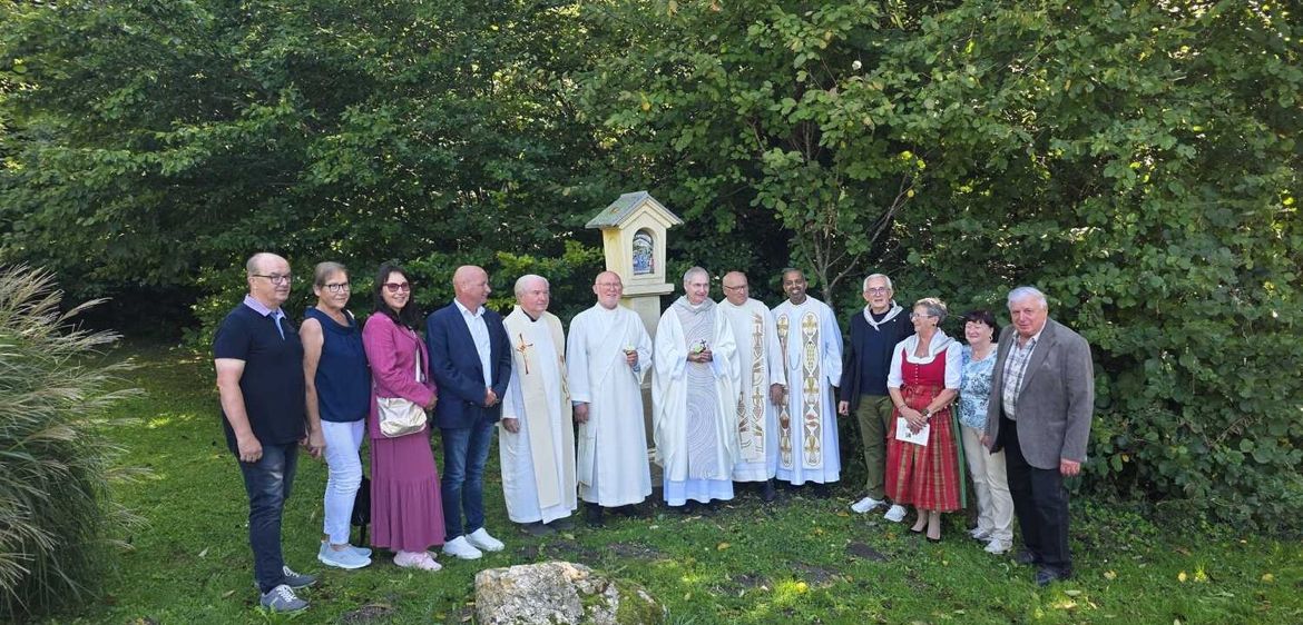 Eine Gruppe von Menschen, einschließlich Geistlicher und Laien, steht draußen in einem grünen Bereich, mit Bäumen und einer kleinen Kapelle im Hintergrund.