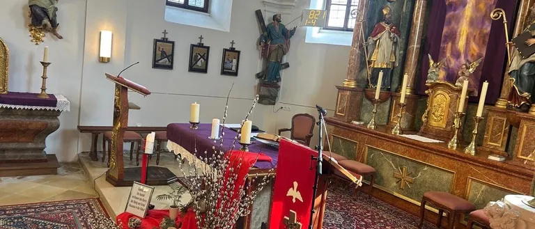 Eine Kircheninnenansicht mit einem Tisch und einer Blumenvase darauf. Eine Statue eines Heiligen steht rechts und ein Kruzifix hängt an der Wand.