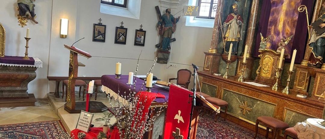 Eine Kircheninnenansicht mit einem Tisch und einer Blumenvase darauf. Eine Statue eines Heiligen steht rechts und ein Kruzifix hängt an der Wand.