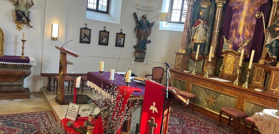 Eine Kircheninnenansicht mit einem Tisch und einer Blumenvase darauf. Eine Statue eines Heiligen steht rechts und ein Kruzifix hängt an der Wand.