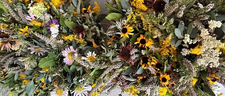 Eine Nahaufnahme von zwei kreisförmigen Blumenarrangements, bestehend aus einer Mischung von Sonnenblumen, Gänseblümchen und verschiedenen grünen Blättern, auf einem grauen Hintergrund angeordnet.