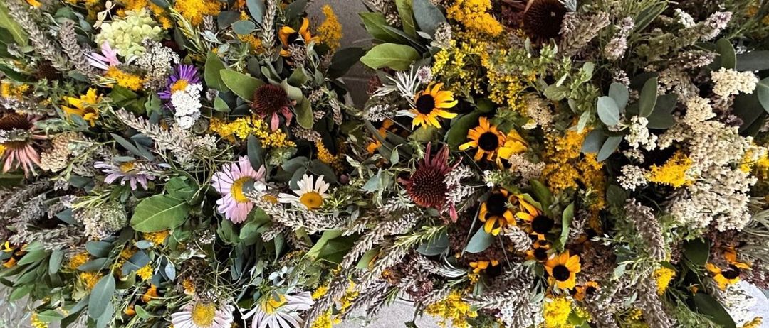 Eine Nahaufnahme von zwei kreisförmigen Blumenarrangements, bestehend aus einer Mischung von Sonnenblumen, Gänseblümchen und verschiedenen grünen Blättern, auf einem grauen Hintergrund angeordnet.