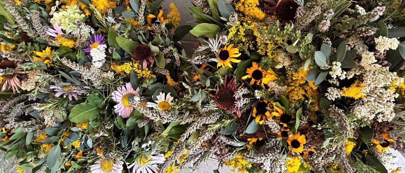 Eine Nahaufnahme von zwei kreisförmigen Blumenarrangements, bestehend aus einer Mischung von Sonnenblumen, Gänseblümchen und verschiedenen grünen Blättern, auf einem grauen Hintergrund angeordnet.