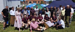 Eine Gruppe von Menschen in traditioneller Kleidung posiert für ein Foto auf einer Rasenfläche mit Zelten und Gebäuden im Hintergrund.
