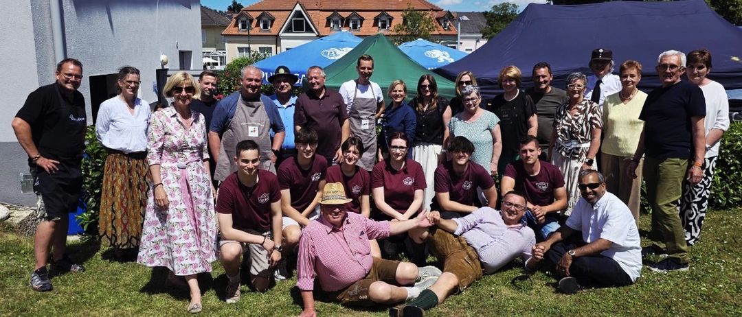 Eine Gruppe von Menschen in traditioneller Kleidung posiert für ein Foto auf einer Rasenfläche mit Zelten und Gebäuden im Hintergrund.