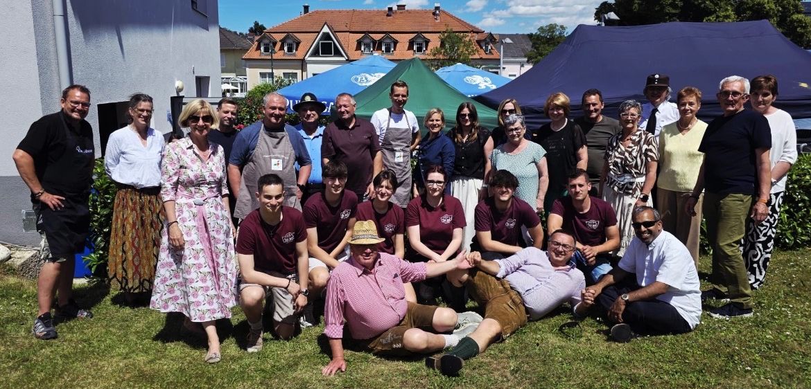Eine Gruppe von Menschen in traditioneller Kleidung posiert für ein Foto auf einer Rasenfläche mit Zelten und Gebäuden im Hintergrund.