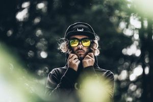Ein Mann mit schwarzer Jacke, Hut und gelber Sonnenbrille steht draußen und hat die Hände am Kinn.