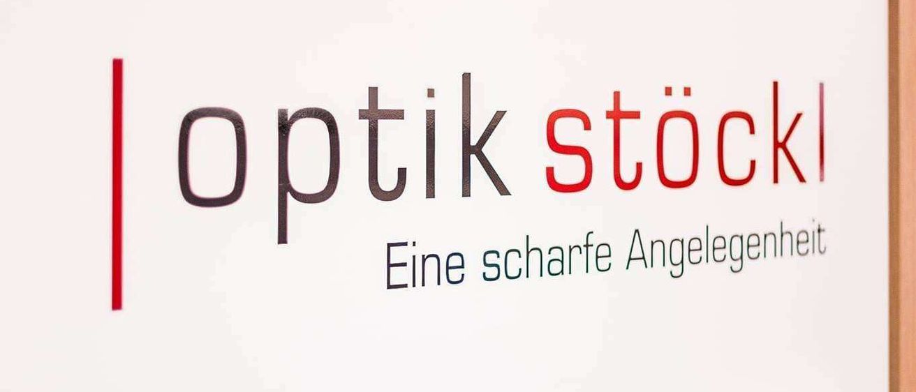 Ein weißes Schild mit den Worten 'Optik Stoeckl' in schwarz und rot, was 'scharfes Sehen' bedeutet, steht vor einem hölzernen Hintergrund.