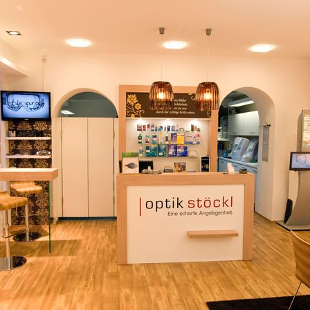 Innenbereich eines Optikgeschäfts. Eine Theke mit der Aufschrift 'Optik Stöckl' steht im Vordergrund. Hinter ihr sind Regale mit Produkten zu sehen. Ein Monitor zeigt die Worte 'style your eye' an.