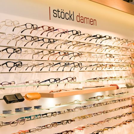 Eine Wand mit einer Vielzahl von Brillen, die auf Regalen ausgestellt sind. Das Schild darüber lautet 'stoeckl damen'. Jede Reihe hat verschiedene Farben und Stile von Brillen.