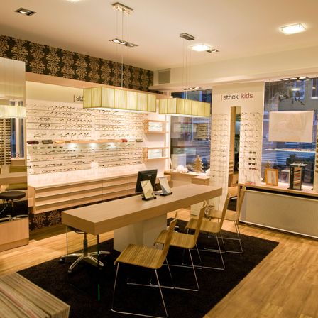 Innenbereich eines Optikerladens mit einem langen Schreibtisch, Holzstühlen, hängenden Lampen, Regalen mit Brillen und einem schwarzen Teppich auf einem Holzboden.