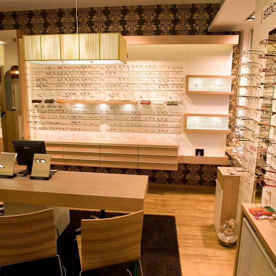 Ein moderner Optikladen mit Holzmöbeln, Regalen mit ausgestellten Brillen, einem Monitor und dekorativen Gegenständen. Der Raum hat einen Holzboden und gemusterte Tapeten.