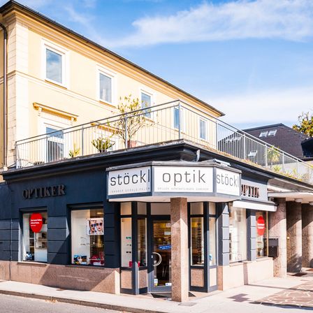 Ein Optikergeschäft namens Optiker steht vor einem großen Gebäude mit Balkon. Es hat ein Schild, das Stockl Optik liest, und eine Topfpflanze auf dem Balkon.
