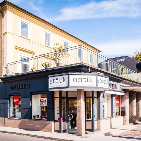 Ein Optikergeschäft namens Optiker steht vor einem großen Gebäude mit Balkon. Es hat ein Schild, das Stockl Optik liest, und eine Topfpflanze auf dem Balkon.