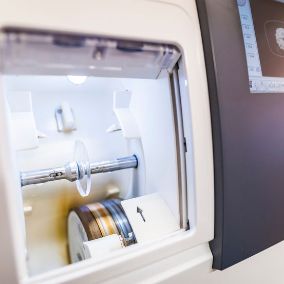 Eine Nahaufnahme einer dentalen Fräsmaschine mit offener Kammer und einem Metallstab im Inneren. Die Kammer ist beleuchtet und hat einen Monitor an der Seite.