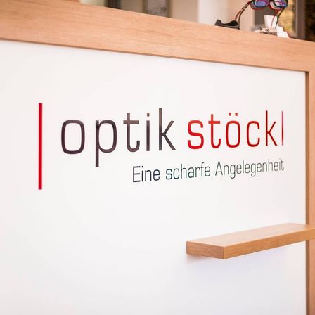 Ein weißes Schild mit 'Optik Stockl' in fetten, schwarzen und roten Schriften. Darunter steht ein Slogan auf Deutsch, 'Eine scharfe Angelegenheit.' Ein hölzerner Rahmen umgibt das Schild.