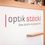 Optik Stöckl-Logo. Ein weißes Schild mit 'Optik Stockl' in fetten, schwarzen und roten Schriften. Darunter steht ein Slogan auf Deutsch, 'Eine scharfe Angelegenheit.' Ein hölzerner Rahmen umgibt das Schild.