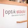 Ein weißes Schild mit 'Optik Stockl' in fetten, schwarzen und roten Schriften. Darunter steht ein Slogan auf Deutsch, 'Eine scharfe Angelegenheit.' Ein hölzerner Rahmen umgibt das Schild.