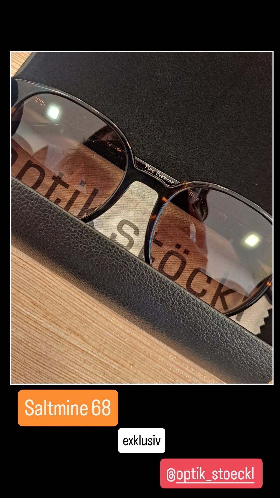 Ein Nahaufnahme von einer Sonnenbrille mit 'optik S' und 'LOCK' Branding, reflektiert auf den Gläsern. Die Gläser sind klar und der Rahmen ist schwarz.