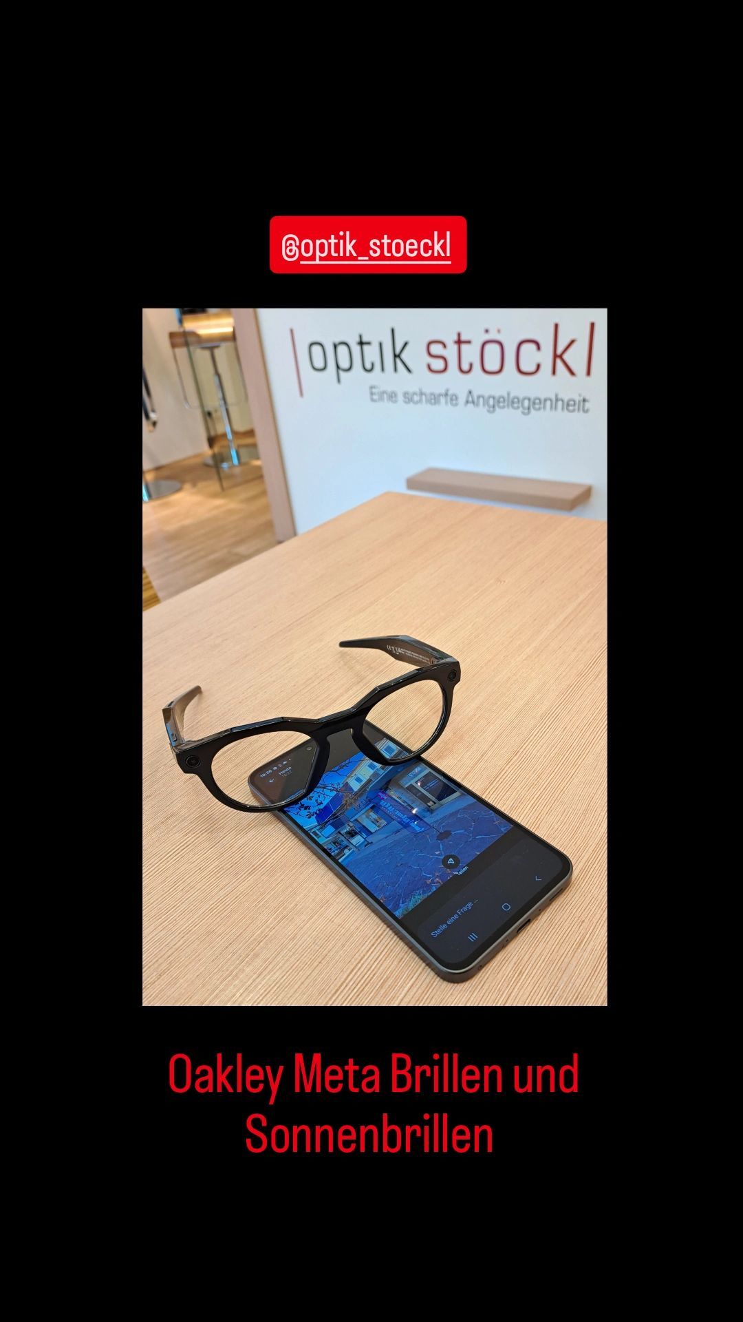 Ein Paar schwarze Brille liegt auf einem Smartphone, das eine Kartenoberfläche anzeigt, auf einem Holztisch in einer Optiker-Filiale.