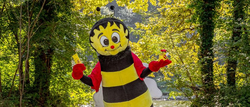Eine Person in einem Bienen-Mascot-Kostüm steht auf einer Holzbrücke, umgeben von Bäumen. Sie hält ein gelbes Objekt in einer Hand und ein rotes Objekt in der anderen.