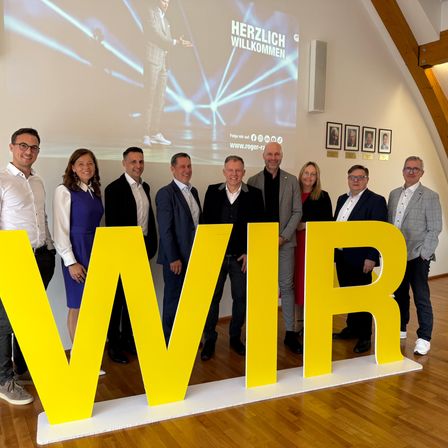 Eine Gruppe von Menschen in formeller Kleidung steht hinter einem großen gelben Schild mit den Buchstaben 'WIR'. Im Hintergrund befindet sich eine Projektionsfläche mit einem Mann in Anzug und Brille.