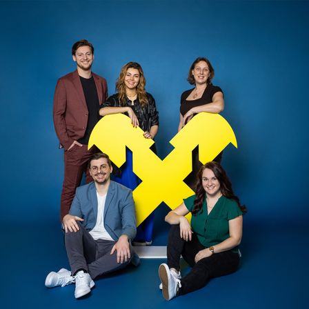 Fünf Personen, vier Frauen und ein Mann, posieren für ein Foto vor einem blauen Hintergrund mit einem großen gelben X-Symbol.