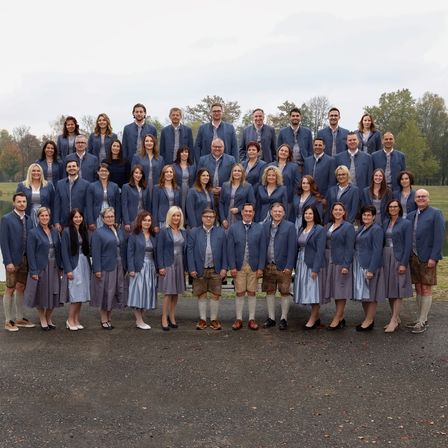Eine große Gruppe von Menschen, die in passenden blauen Blazern und Röcken oder Kleidern gekleidet sind, posiert für ein Foto vor einer ruhigen, von Bäumen gesäumten Kulisse.