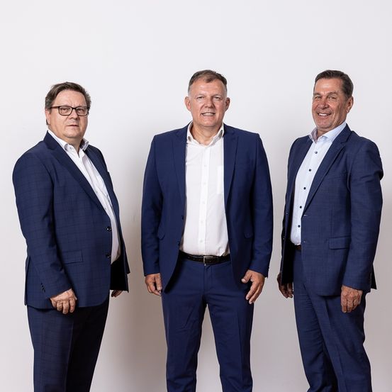 Drei Männer in Anzügen stehen nebeneinander vor einem weißen Hintergrund und lächeln für die Kamera. Der Mann auf der linken Seite trägt eine Brille.
