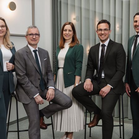 Fünf Fachleute in Business-Kleidung, darunter zwei Frauen, posieren für ein Foto in einer modernen Büroumgebung.