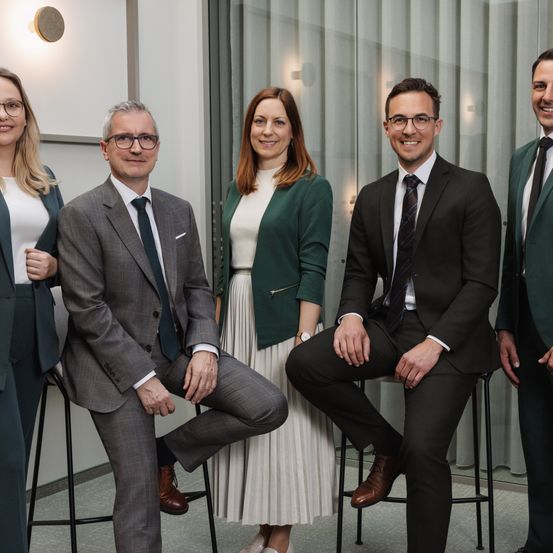 Fünf Fachleute in Business-Kleidung, darunter zwei Frauen, posieren für ein Foto in einer modernen Büroumgebung.