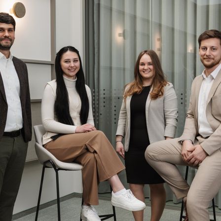 Vier Menschen in professioneller Kleidung stehen drinnen zusammen. Eine Frau in brauner Hose und einem weißen Rollkragenpullover sitzt auf einem Stuhl. Eine Frau in einem schwarzen Oberteil und einer grauen Jacke steht neben ihr. Ein Mann in einem Anzug steht hinter ihnen.