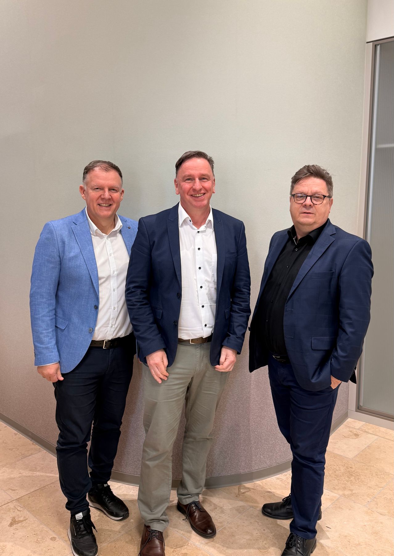 Drei Männer in Anzügen stehen nebeneinander in einem Büro. Der Mann auf der linken Seite trägt einen hellblauen Blazer. Der Mann in der Mitte trägt einen dunkelblauen Blazer und ein weißes Hemd. Der Mann auf der rechten Seite trägt einen dunkelblauen Blazer und ein schwarzes Hemd.