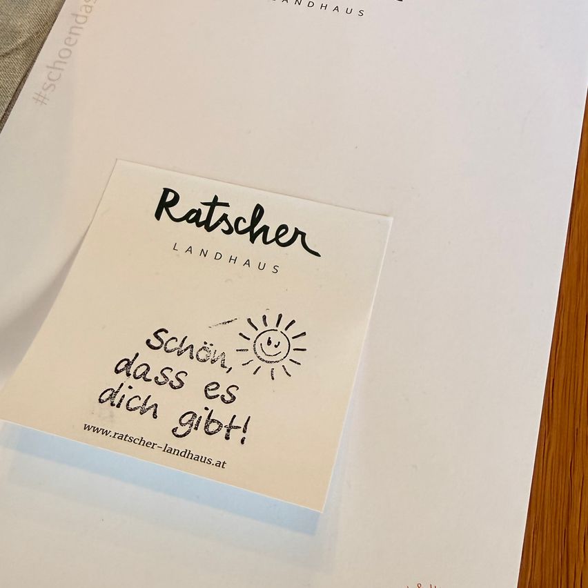 A white paper with the text 'Ratscher Landhaus' has a small note on it that says 'Schön, dass es dich gibt!' and includes a sun drawing.