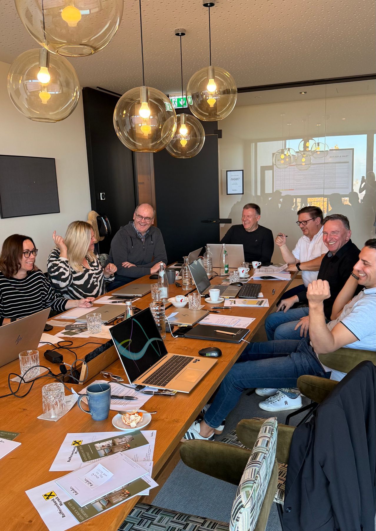 A group of people sit around a table with laptops and cups, smiling and talking. The table is filled with papers and a bowl of food.
