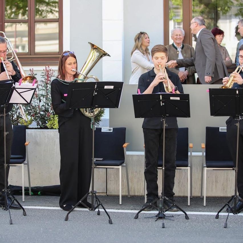 Eine Gruppe Musiker spielt Blechblasinstrumente auf einer Bühne vor einem Gebäude. Einige Leute schauen zu.