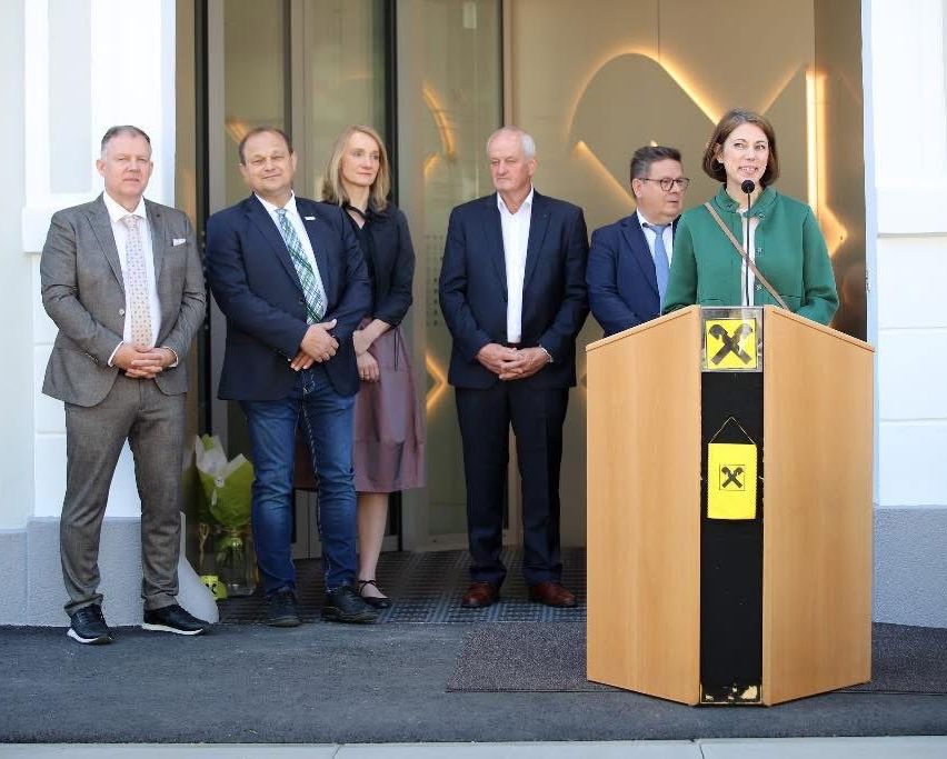 Fünf Menschen stehen vor einem Podium mit einem gelben Schild mit einem schwarzen X. Sie sind alle in Anzug und Krawatte gekleidet.