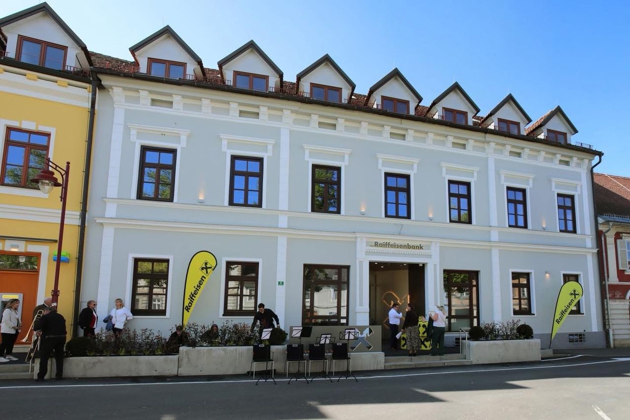 Ein weißes Gebäude mit mehreren Fenstern, einem gelben Banner auf der linken Seite und einem Schild, das Raiffeisenbank besagt. Leute sind draußen und Stühle und Notenständer sind vor dem Gebäude aufgestellt.