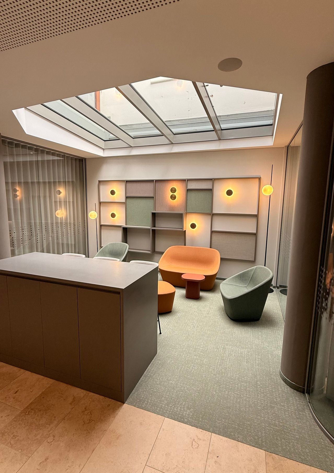 Ein Innenraum mit Glasdecke. Ein Sofa, Stühle und ein Schreibtisch stehen auf dem Boden. Eine Lampe steht auf der rechten Seite des Raumes.