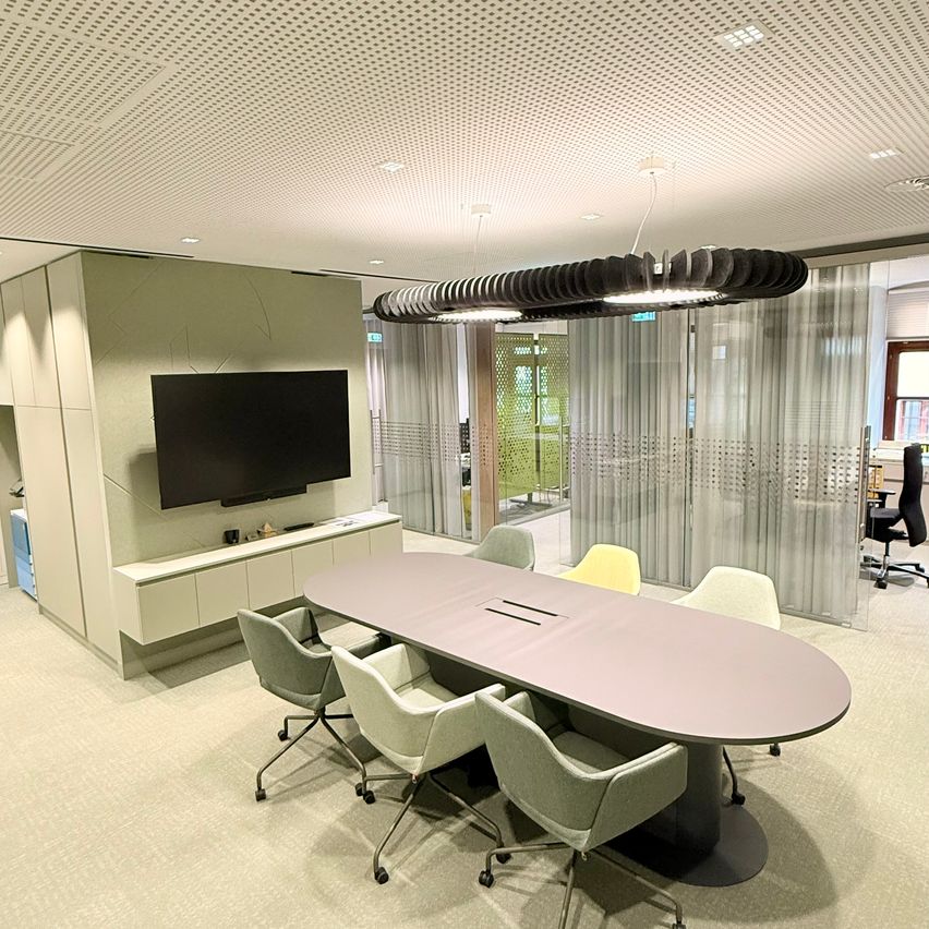 Ein moderner Bürobesprechungsraum mit einem großen Tisch, Stühlen und einem Fernseher. Der Raum hat eine Decke mit Lichtern und weißen Wänden.