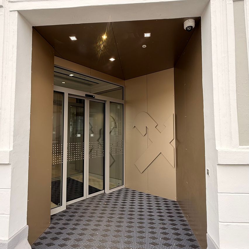 Ein leerer Eingangsflur mit Glastüren, gefliestem Boden und braunen Wänden, ausgestattet mit einer Überwachungskamera und einem Muster an einer Wand.