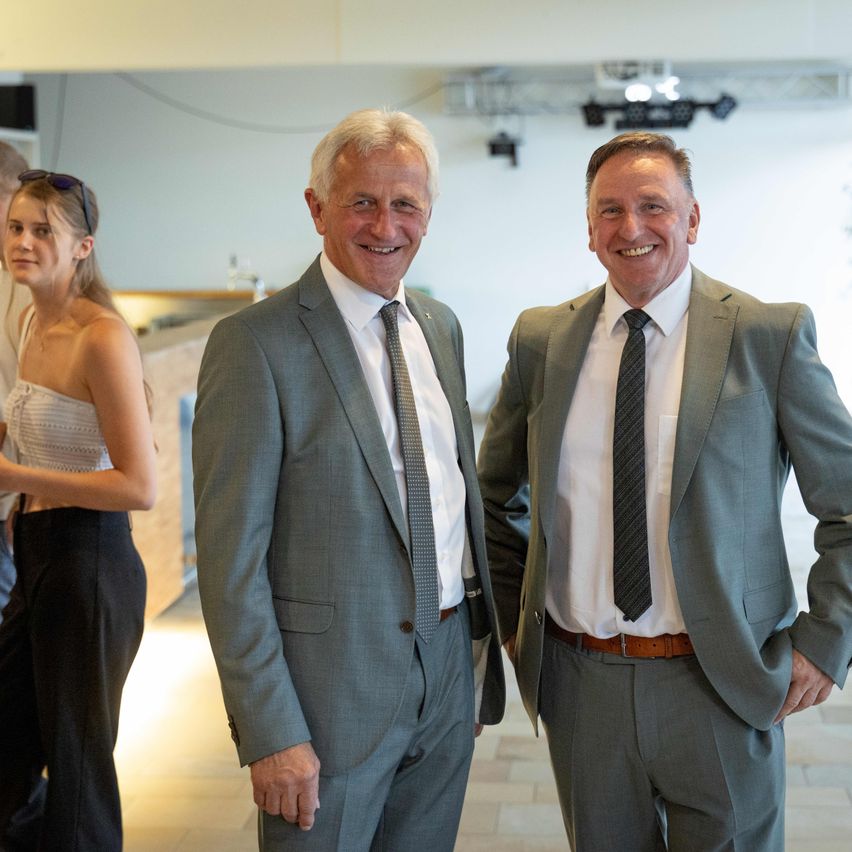 Two men in suits stand smiling with a woman in casual attire in a bright room. The room has white walls and tiled floors, with a white shelf in the background.