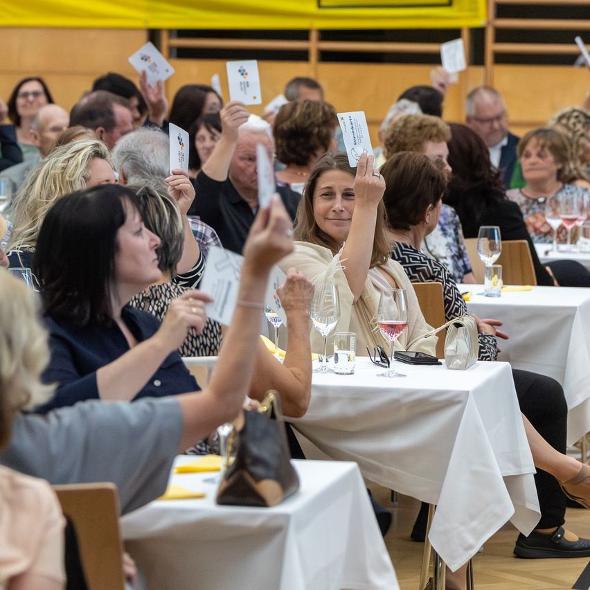 A group of people in a conference hall are holding up cards. Some are sitting at tables with wine glasses and white tablecloths. The woman in the middle is smiling and raising her card.
