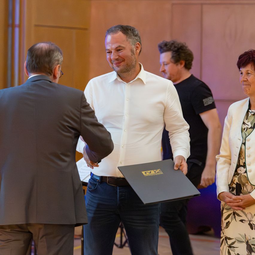 A man in a white shirt holds a folder and smiles. Another man in a suit looks at him. A woman in a floral dress stands nearby. A man in a black shirt stands behind them.