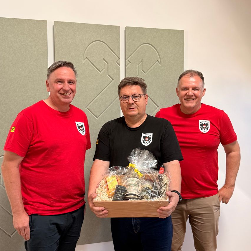 Drei Männer in roten T-Shirts posieren mit einer Geschenkbox voller Lebensmittel. Sie stehen vor einer Kulisse mit großen Buchstaben.