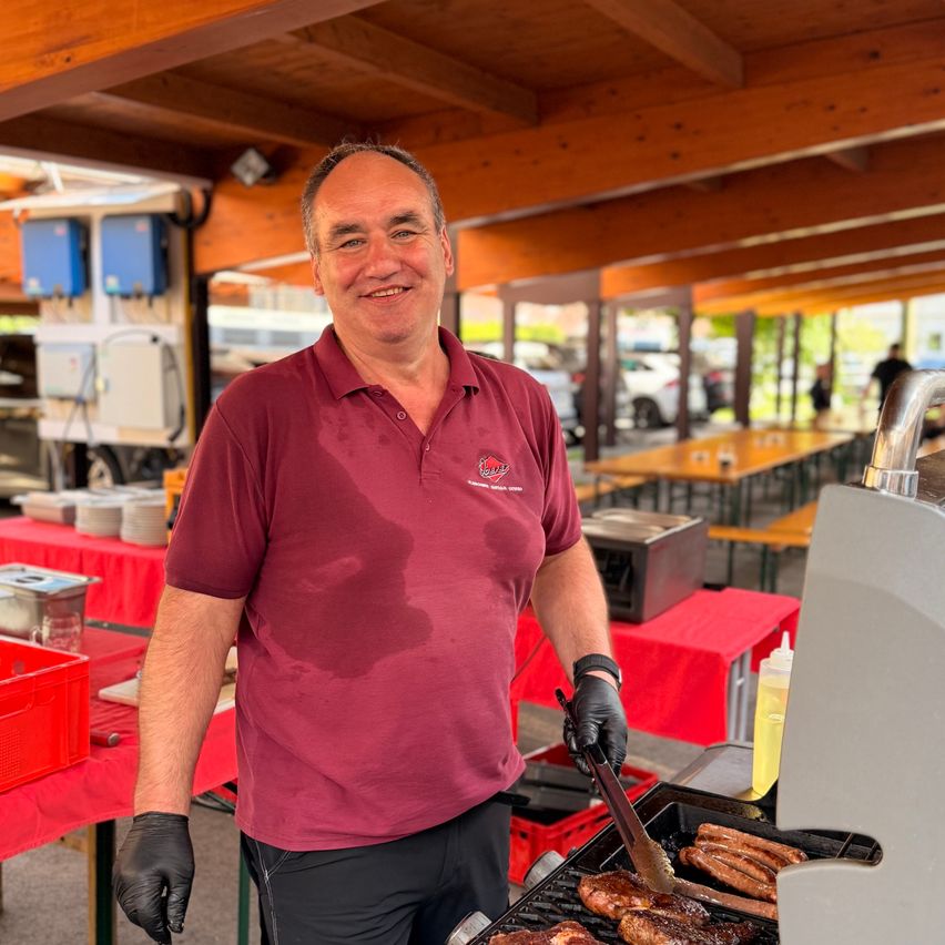 Ein Mann in einem weinroten Poloshirt und schwarzen Handschuhen grillt Würste und Fleisch auf einem Grill bei einer Outdoor-Veranstaltung unter einer Holzkonstruktion.