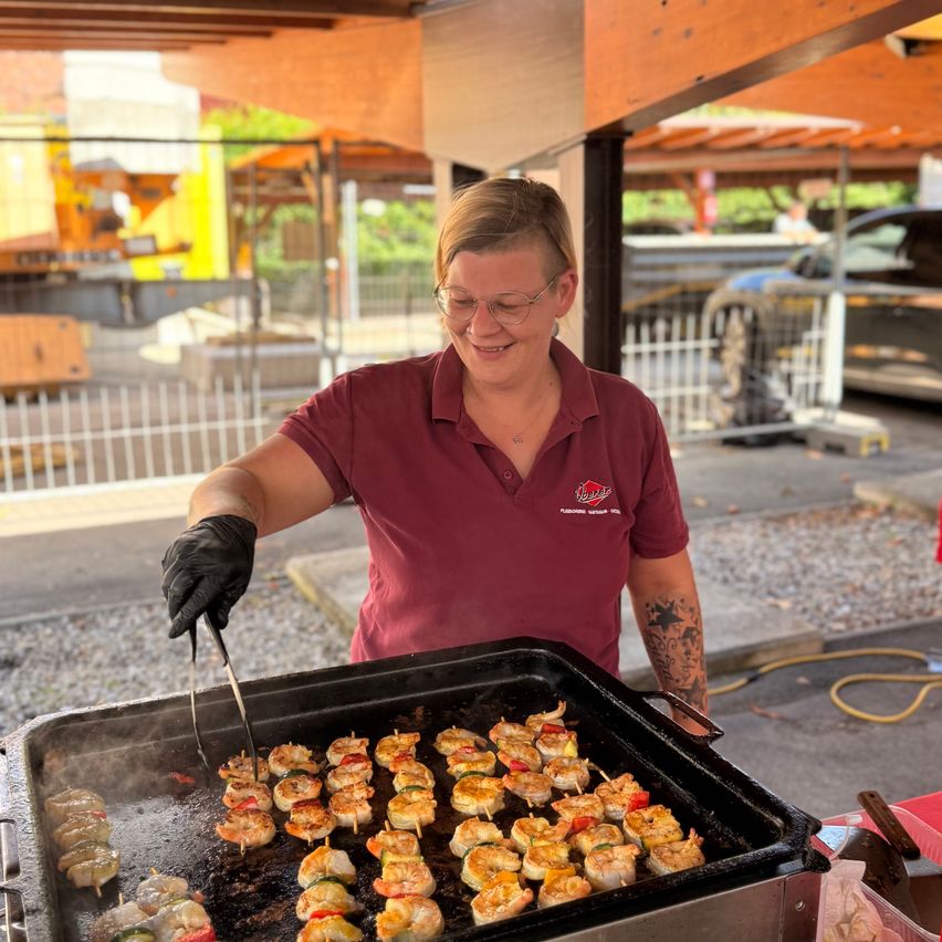 Eine Frau in einem weinroten Shirt und schwarzen Handschuhen grillt Essen auf einem Grill im Freien.
