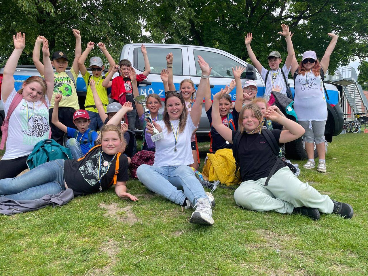Eine Gruppe von Kindern und Erwachsenen feiert auf einem Rasenfeld mit einem Van im Hintergrund. Einige sitzen und andere stehen mit erhobenen Händen. Ein Kind hält einen Pokal.